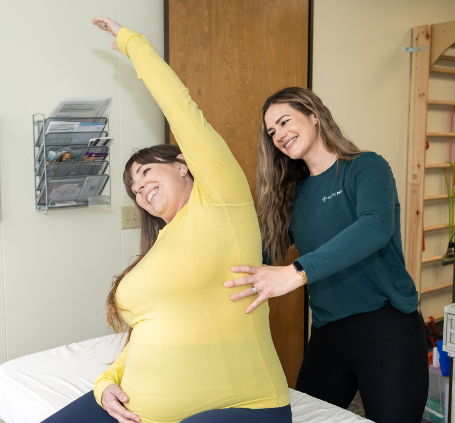 Two women in a medical or therapy setting, one assisting the other with a stretch.